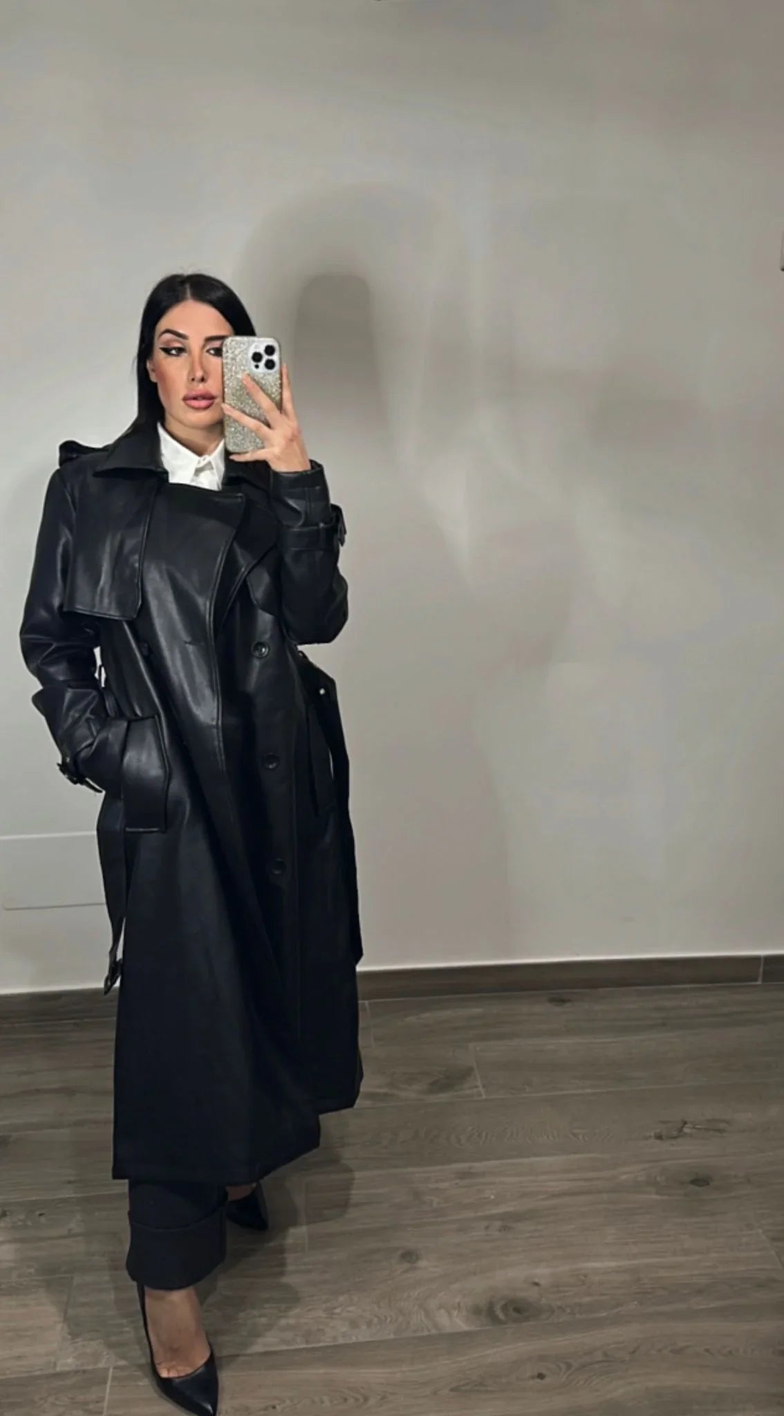 Donna con trench nero ecologico lucido, camicia bianca e tacchi neri, stile moda elegante.
