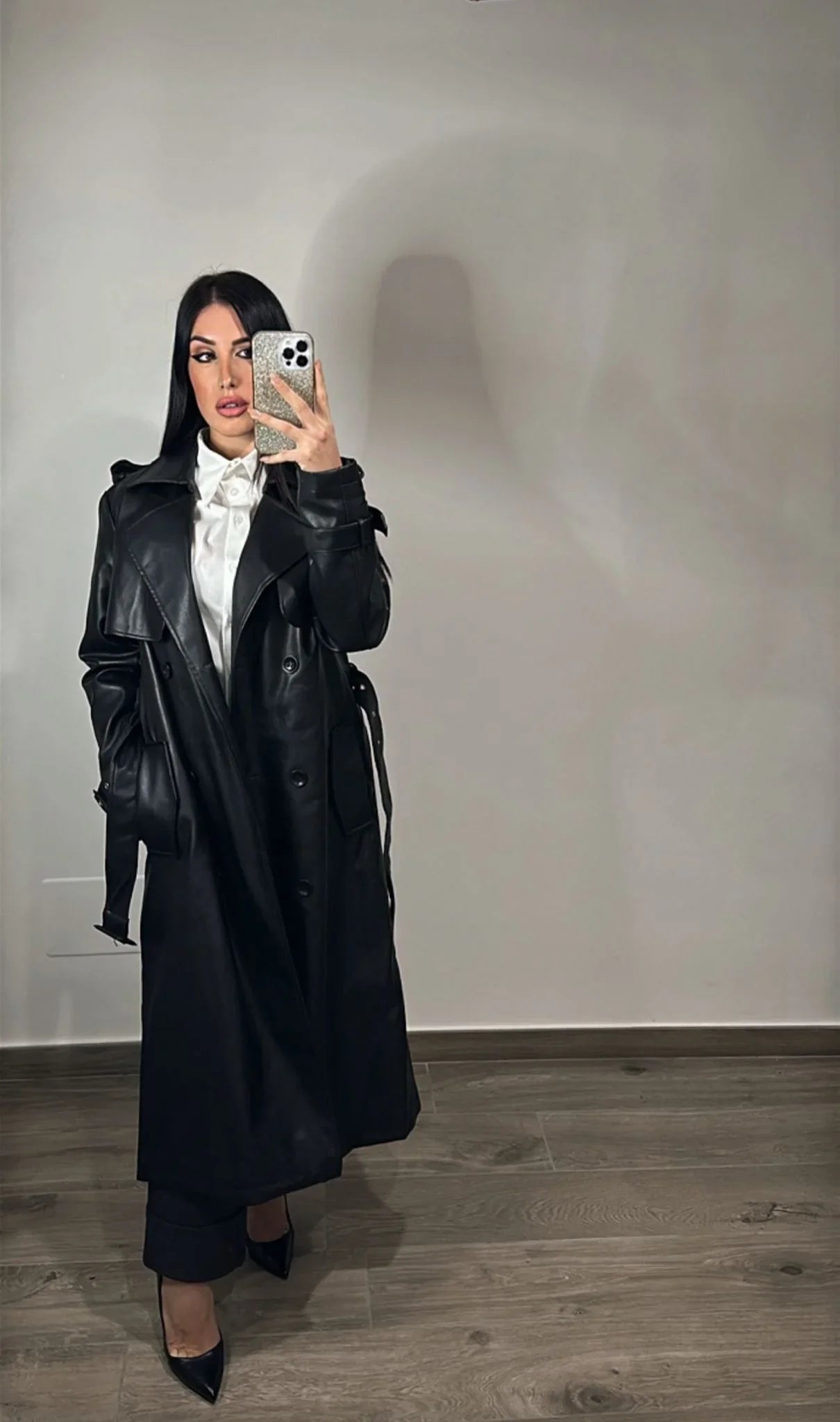 Donna indossa trench nero eco pelle, camicia bianca e tacchi, posa su pavimento in legno