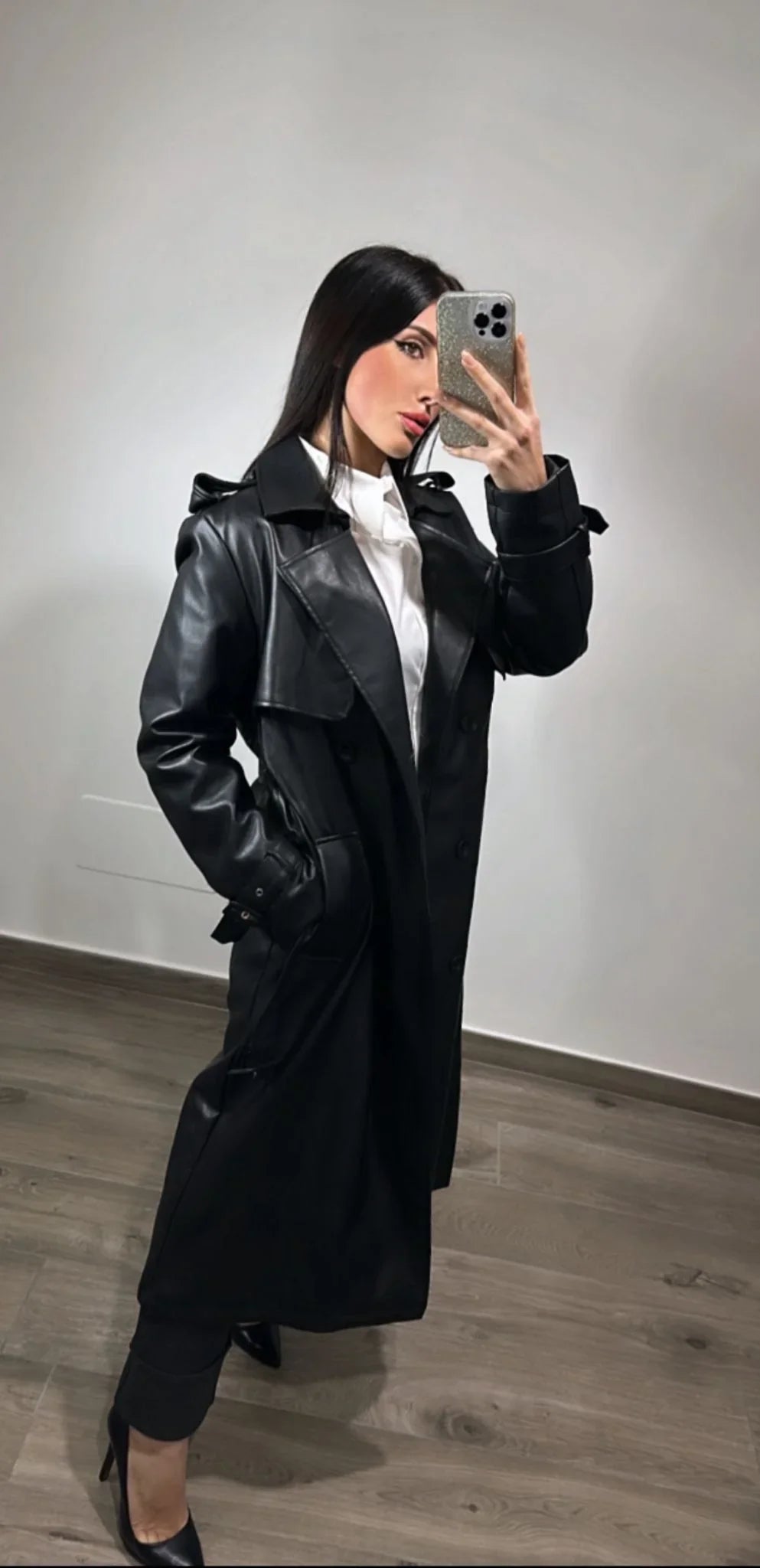 Donna con trench nero ecopelle, camicia bianca e tacchi in una stanza minimal.