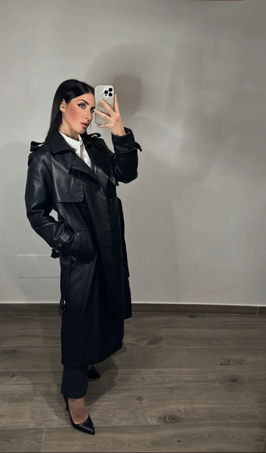 Donna con trench nero elegante in ecopelle, camicia bianca e tacchi in un ambiente moderno